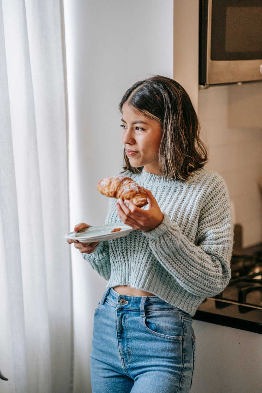 smiling ethnic woman eating croissant in kitchen